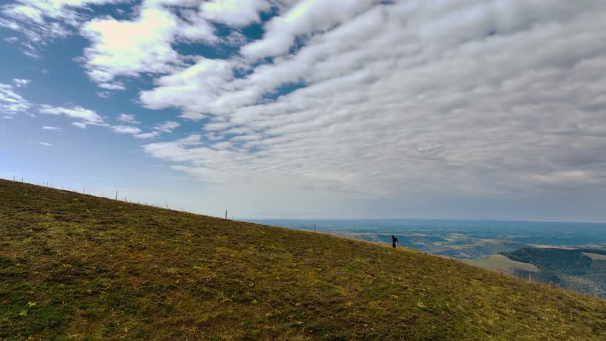 Drone aerial perspective showing a lone photographer standing on a wide grassy hillside. Expansive landscape under a textured cloudy sky.