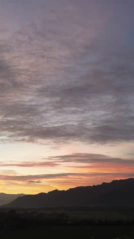 Aerial drone view of rural farmland at sunset with dramatic clouds and mountain silhouettes. Warm evening sky creates a calm and atmospheric countryside landscape.