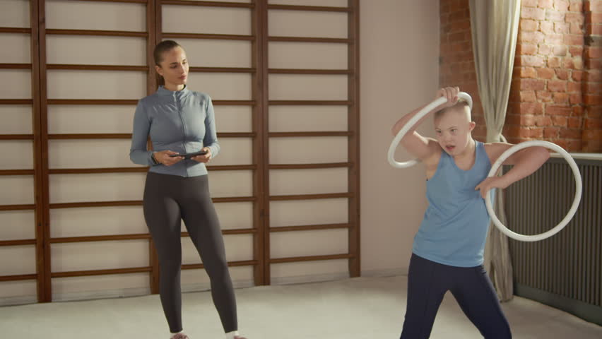 Young boy with down syndrome practicing physical exercise using hoop while female trainer supervising during inclusive gymnastics class focused on coordination and motor skills