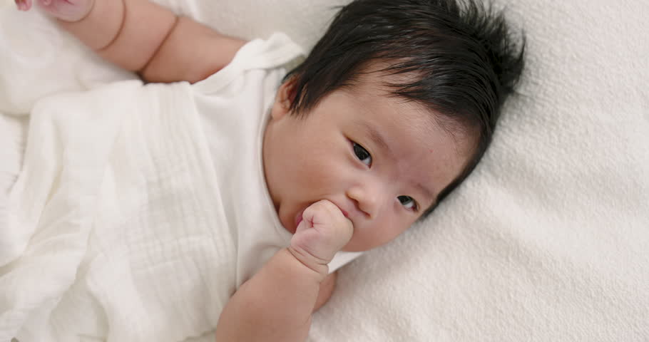 Asian infant boy lying on bed wrapped in white blanket sucking thumb with soft expression showing self-soothing and comfort during early bonding babycare time