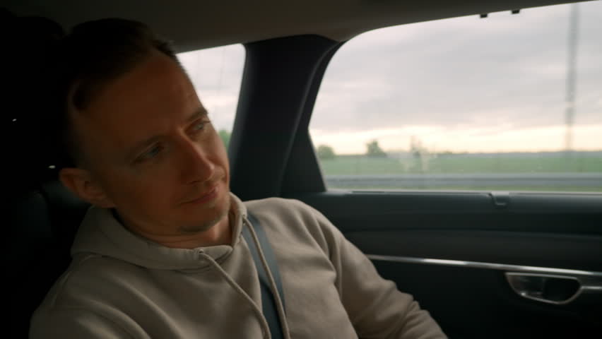Male Passenger Gazing Out Window At Passing Fields And Sunset, Calm Expression, Seatbelt On And Hoodie