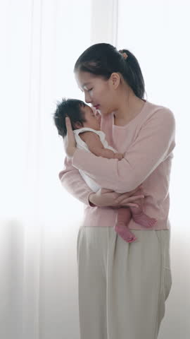 Asian mother holding newborn baby boy near window, leaning in to kiss head softly, expressing emotional closeness and tenderness during peaceful morning bonding moment indoors