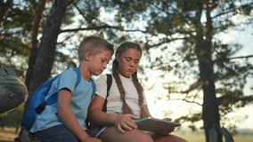 Girl shows screen to boy in forest. Children sit on fallen tree using tablet. Outdoor sibling learning experience. Forest tablet children sit. Device in hands of children during outdoor break. - Powered by Shutterstock - Get 15% off with code: PIKWIZARD15