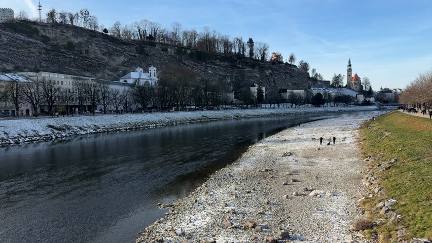 People walking along the Salzach River in Salzburg during winter. Calm handheld shot with sunlight, showing bare trees, riverside paths, and historic architecture against a rocky hillside