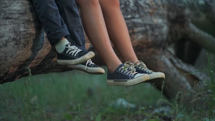 Children feet in sneakers on log. Boy rests feet while sitting. Forest trees around children sitting. Sneakers hang below feet. Children sneakers worn on sitting log. Forest calm with feet down.