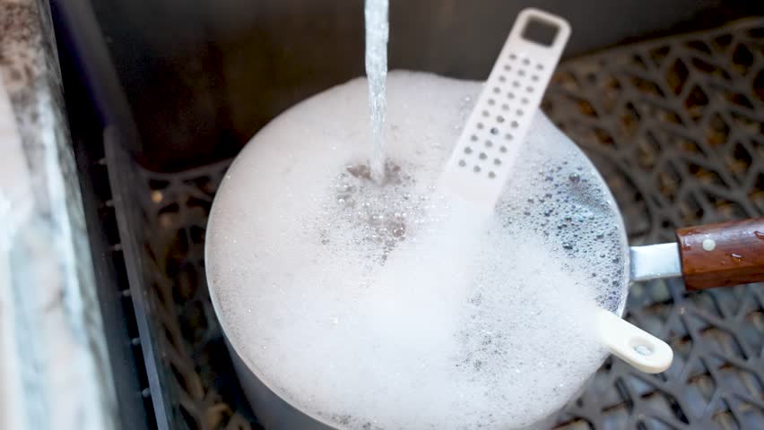 A pot filled with soap suds sits in a kitchen sink under running water ready for cleaning.