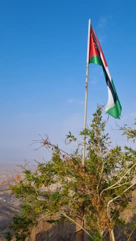 Vertical shot of a Jordanian flag fluttering lightly against a clear blue sky on a mountain overlooking Wadi Araba, with the vast desert landscape stretching below