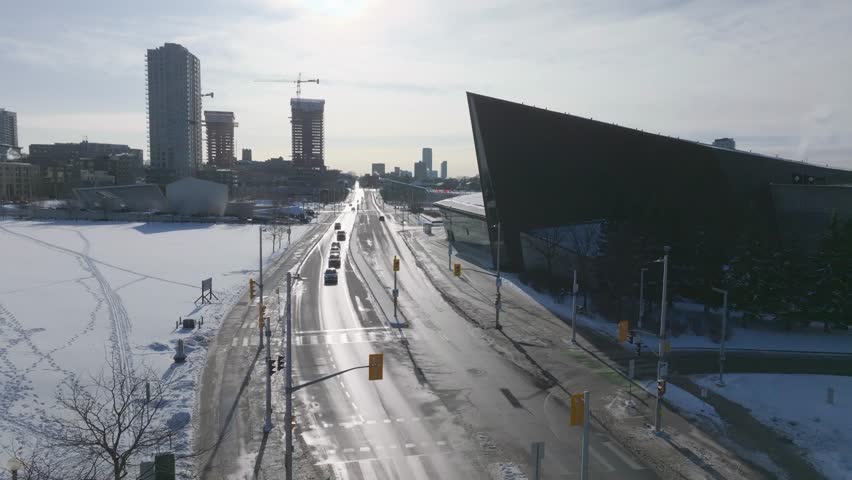 Aerial view of the Canadian War Museum in Ottawa on a sunny winter day. Traffic moves along the snowy road near LeBreton Flats with construction cranes and city skyline in the background.