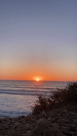 beautiful sunset over a calm ocean with gentle waves reaching the sandy beach, creating a peaceful and warm evening atmosphere with soft orange and pink sky tones.