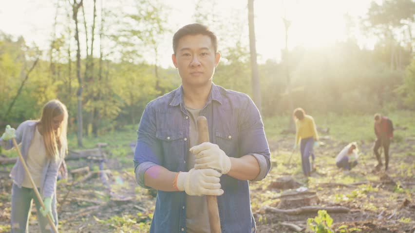 Front view of Asian man holding shovel handle. Young male wearing gloves during tree planting. Group of volunteers working behind him. Reforestation activity in forest. Environmental reforestation.