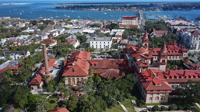 Historic red-roofed architecture dominates the scene near the waterfront with boats anchored in the bay and a long bridge connecting to the mainland in St Augustine, Florida.