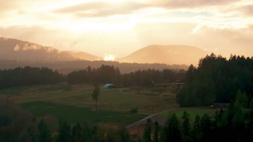Aerial footage of a drone rising over forested hills and farmland as warm sunset light and drifting fog surround the mountains in Cowichan Valley, British Columbia.
