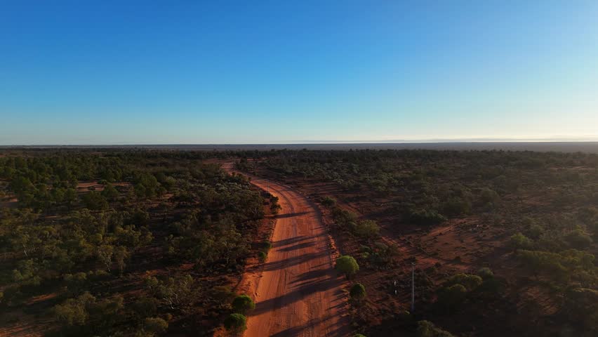 Aerial of remote New South Wales outback with dirt road cutting through vast arid landscape under clear sky, Australian wilderness