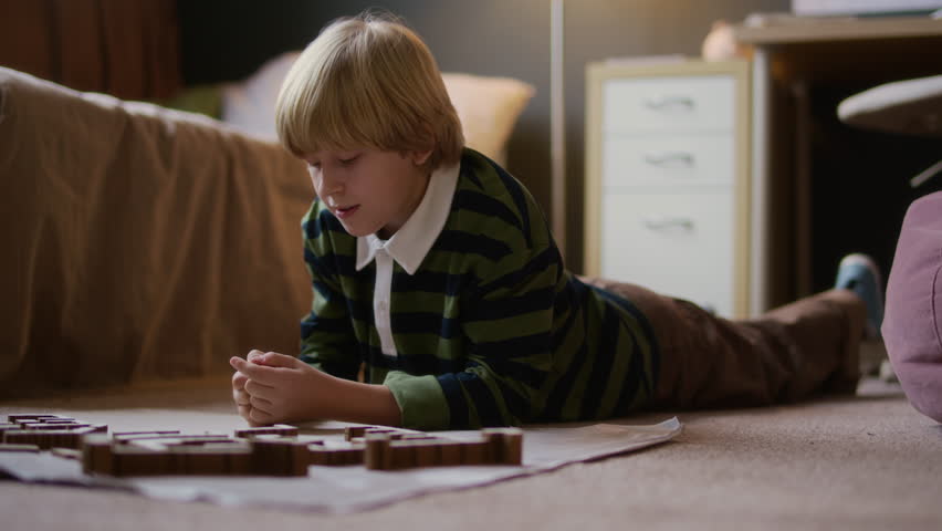 Ground level slowmo shot of inspired blond Caucasian boy child enjoying assembling wooden puzzle blocks while lying on floor in his cozy modern room