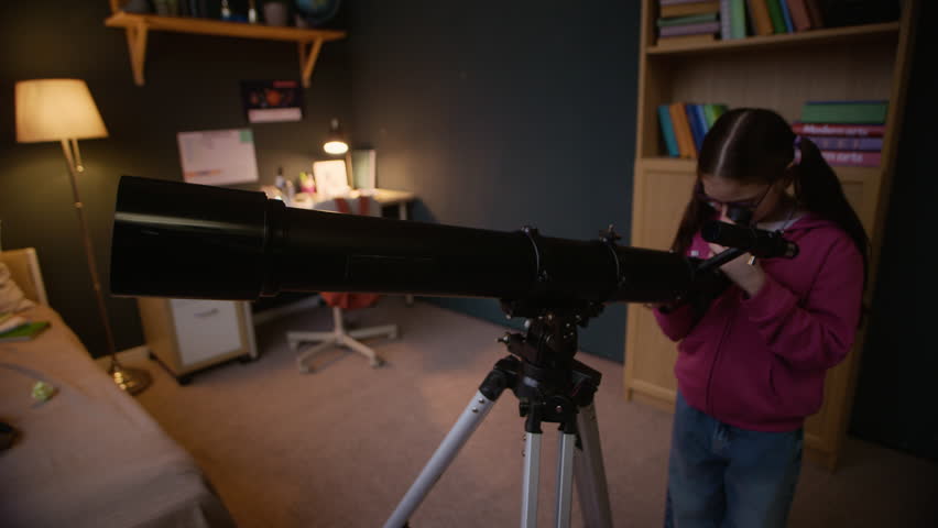 Slowmo shot of Caucasian girl child in eyeglasses using telescope to observe night sky while standing in her cozy room in evening