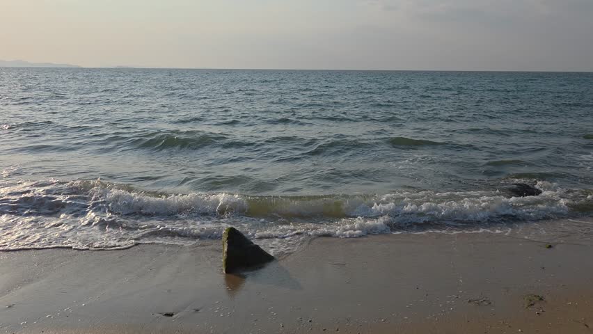 A wide, eye-level shot of a sandy beach with a single dark rock in the foreground as gentle waves wash ashore under a clear sky.	
