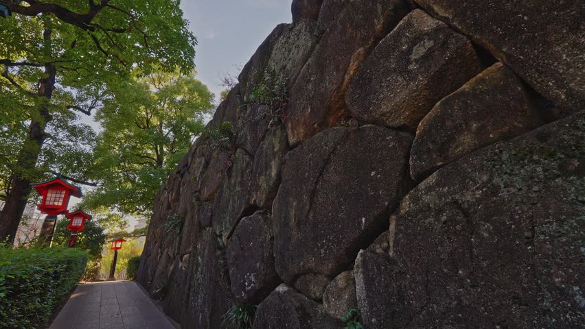 A perspective shot of a massive, ancient stone fortification wall bordering a garden path lined with traditional red lanterns.