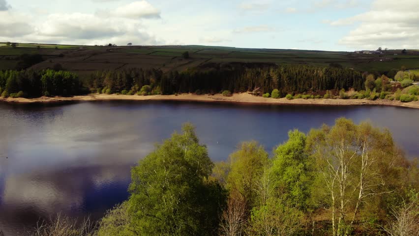 Trees and farmland surround lake in Peak District National Park blue sky landscape wide 4k drone aerial selective focus