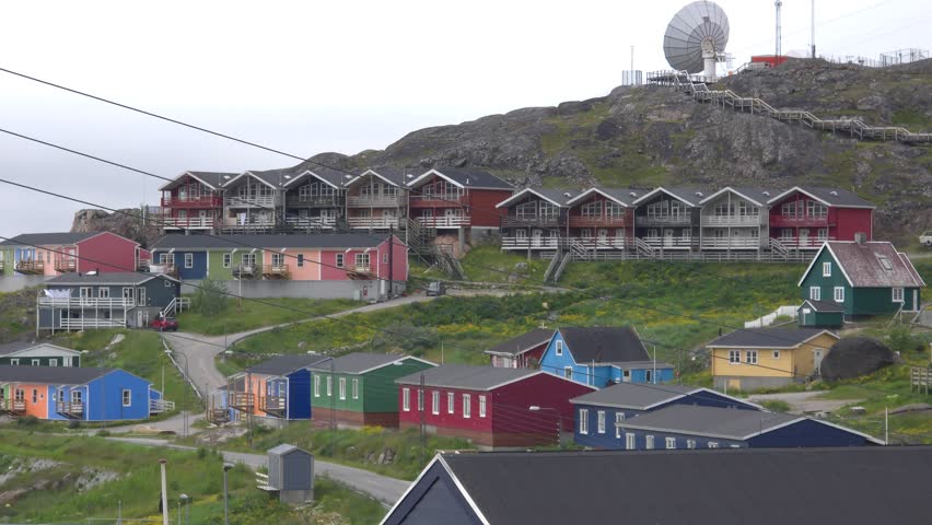 A panoramic view of a settlement with numerous colorful houses built on a steep hillside. A large satellite dish is visible on the hilltop, overlooking the town. The houses are of various sizes and colors, including red, blue, green, yellow, and pink. Some houses are terraced, with stairs leading up to them. The surrounding landscape is rocky and covered with sparse vegetation. The sky is overcast. Shot on: November 18, 2025 at 12:06 PM. Location: Qaqortoq, Kalaallit Nunaat.Greenland