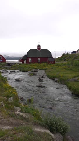 A red building with a dark roof and white-framed windows stands beside a flowing stream. The building has a cross on its steeple. The surrounding area is covered in green grass and small yellow flowers. The scene appears to be in a small town with other buildings visible in the background. Shot on: November 18, 2025 at 12:06 PM. Location: Qaqortoq, Kalaallit Nunaat. Located in Greenland.