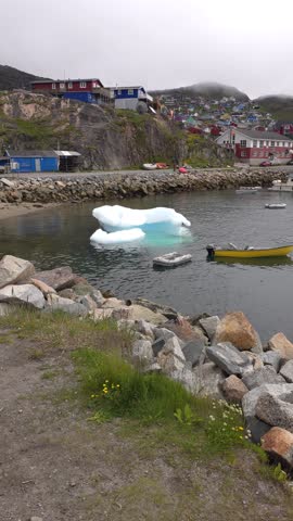 A bright blue and white iceberg floats in a calm harbor, with a town of colorful buildings nestled on the hillside behind it. Boats are docked along a rocky shoreline, and vegetation grows on the embankment in the foreground. The sky is overcast. Shot on: November 18, 2025 at 12:06 PM. Location: Qaqortoq, Kalaallit Nunaat.Greenland