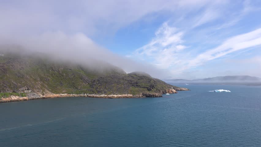 A serene coastal scene unfolds with a rugged, green-covered mountain range partially shrouded in mist. The deep blue ocean stretches towards the horizon, dotted with a few distant icebergs. A small cluster of buildings is visible nestled on the rocky shore. The sky above features wispy clouds against a bright blue backdrop. Shot on: November 18, 2025 at 12:07 PM. Location: Qaqortoq, Kalaallit Nunaat.Greenland