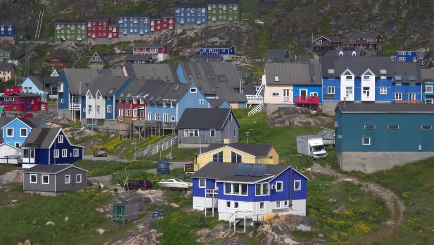 A panoramic view of a town with numerous colorful houses built on a steep, rocky hillside. The houses are painted in various bright colors, including blue, red, yellow, and green, with some featuring white trim. Several cars, a boat, and a white van are visible. The landscape is rugged with green grass and scattered rocks. The scene captures a unique settlement nestled against a natural backdrop. Shot on: November 18, 2025 at 12:07 PM. Location: Qaqortoq, Kalaallit Nunaat.Greenland
