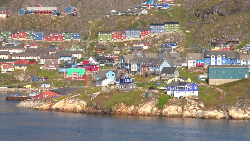 A panoramic view of a coastal town with numerous colorful houses built on a steep, rocky hillside. The houses are painted in vibrant shades of red, blue, green, and yellow, creating a striking contrast against the natural landscape. A body of water is visible in the foreground, with a small dock and some boats. The scene captures the unique architecture and setting of a northern settlement. Shot on: November 18, 2025 at 12:09 PM. Location: Qaqortoq, Kalaallit Nunaat.Greenland