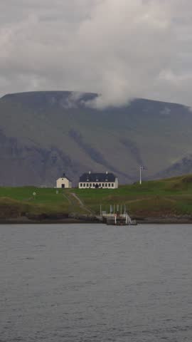 A scenic view of a coastal village nestled against a backdrop of lush green mountains. A large white building with many windows stands prominently, with a smaller white structure nearby. A pier extends into the calm, dark water, with several boats docked. A flag flies on a pole. The sky is overcast with clouds. Shot on: November 18, 2025 at 12:10 PM. Location: Reykjavíkurborg, Iceland.