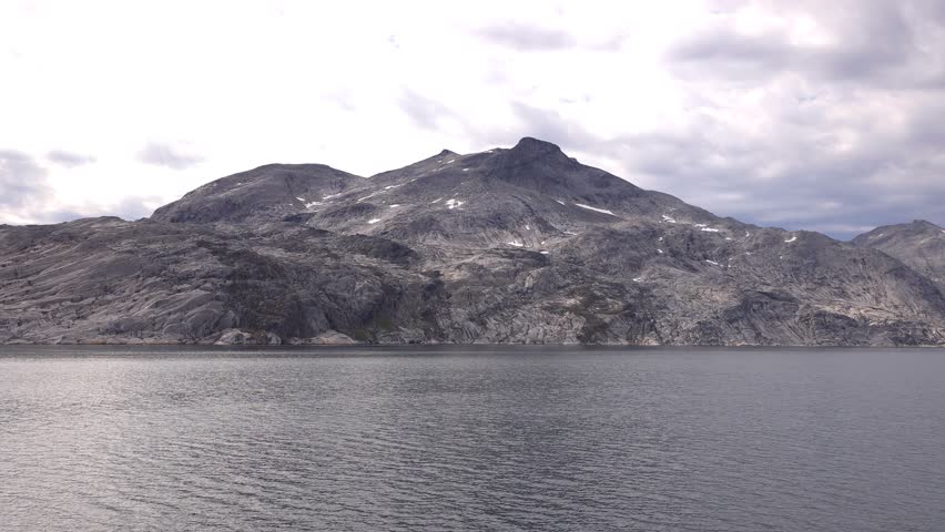 A wide shot captures a vast, calm body of water in the foreground, reflecting the overcast sky. Behind it, a dramatic mountain range dominates the horizon. The mountains are rugged and rocky, with patches of snow or ice visible on their slopes. The overall scene is one of natural grandeur and serene isolation. Shot on: November 18, 2025 at 12:10 PM. Location: Kalaallit Nunaat. Greenland