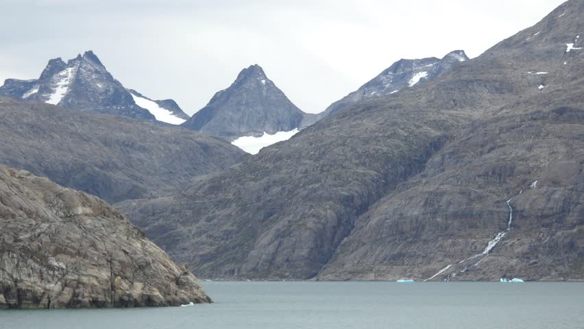 A panoramic view of a serene fjord in Greenland, with towering, rugged mountains dominating the scene. Snow-capped peaks and rocky slopes are visible, with a thin waterfall cascading down one of the mountainsides. The calm, grey water of the fjord reflects the overcast sky, and a few small icebergs are scattered in the distance. The overall impression is one of vast, untouched natural beauty. Shot on: November 18, 2025 at 12:11 PM. Location: Kalaallit Nunaat.Greenland