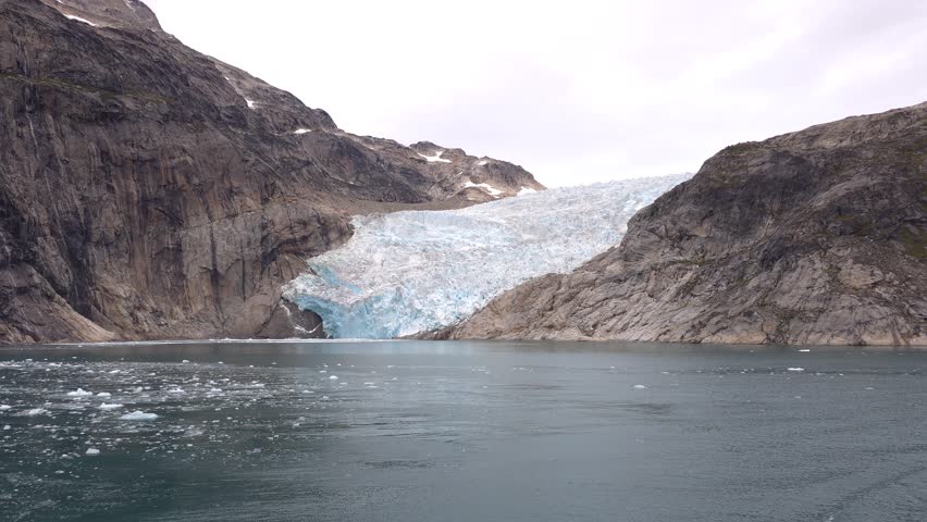 A vast glacier flows down a mountainside, meeting a body of water. The glacier is a brilliant white and blue, with visible crevasses and a large opening at its base. The surrounding mountains are steep and rocky. The water is calm, with small ice floes scattered across its surface. The sky is overcast. Shot on: November 18, 2025 at 12:11 PM. Greenland