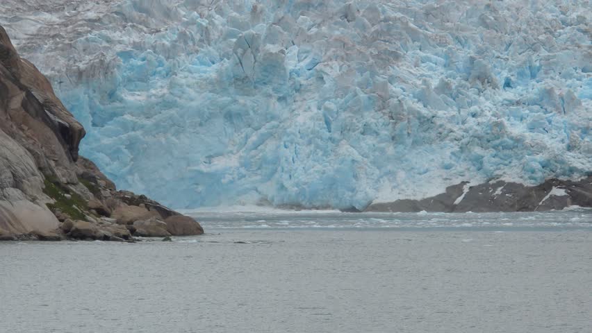 A vast glacier face, tinged with blue hues, meets the water. Ice chunks are visible breaking off and floating in the sea. Rocky cliffs frame the left side of the glacier, with patches of snow on the right. The scene captures the immense power and beauty of glacial landscapes. Shot on: November 18, 2025 at 12:11 PM. Location: Kalaallit Nunaat.Greenland