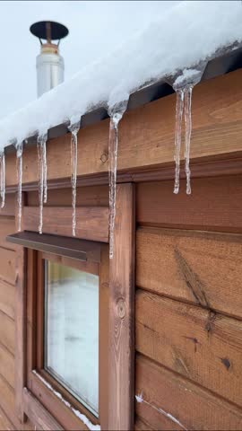 Icicles hang from the roof of a wooden cabin, illustrating the melting snow and ice, with a chimney and wooden siding creating a rustic winter scene