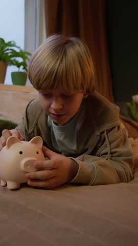 Vertical slowmo shot of dreamy blond Caucasian boy child lying on bed and thinking about his goal while putting coins into piggy bank