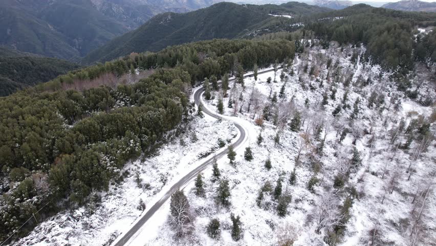 An aerial view of a winding mountain road in a winter forest. A winding road through a snow-covered pine forest under a cloudy sky. A cinematic winter landscape.