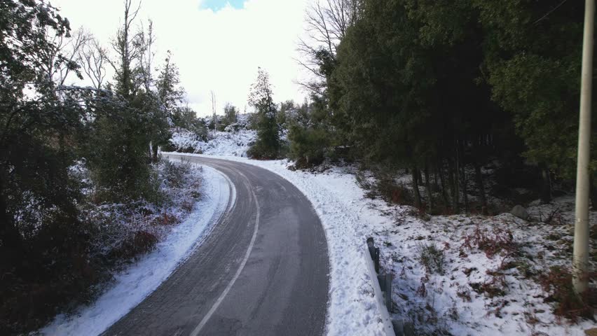 A snow-covered forest road in a winter landscape. A winding asphalt road, covered in snow, winds through a quiet winter forest. Snow-covered trees and a cloudy sky create a cool, peaceful atmosphere.