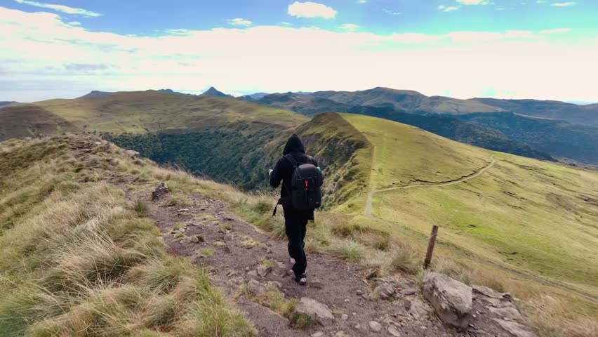 Aerial view of a photographer with a backpack standing on a narrow mountain ridge in Auvergne-Rhône-Alpes capturing photos of the valley.