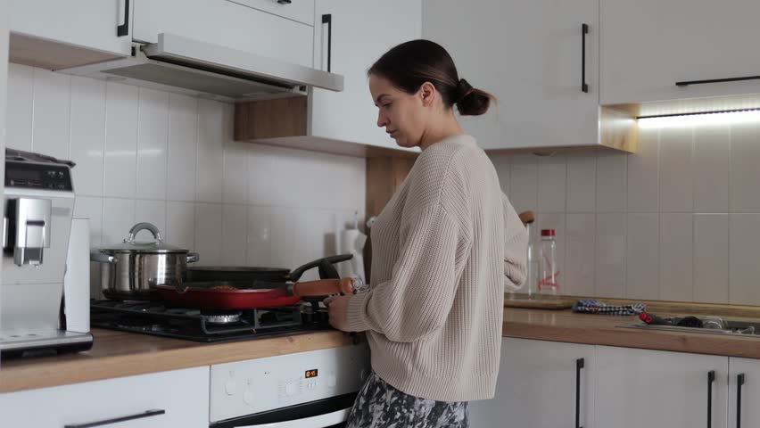 Confident young woman preparing homemade meal, pan-frying ingredients on stovetop within modern domestic kitchen interior