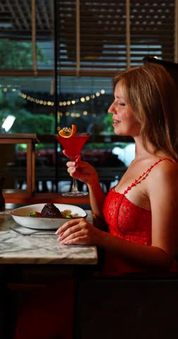 Elegant woman sips red cocktail in dimly lit luxury restaurant. Sophisticated dining experience. Relaxed evening atmosphere. Vertical composition.