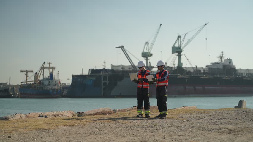 Workers check equipment near ships at a busy port on a sunny day