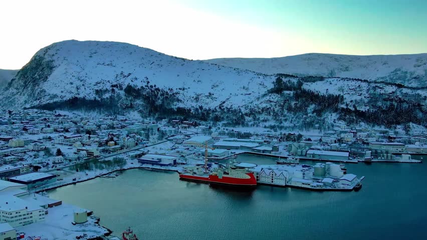 A picturesque winter scene unfolds as a coastal community rests under a blanket of snow, with a vibrant orange vessel moored in the calm harbor waters