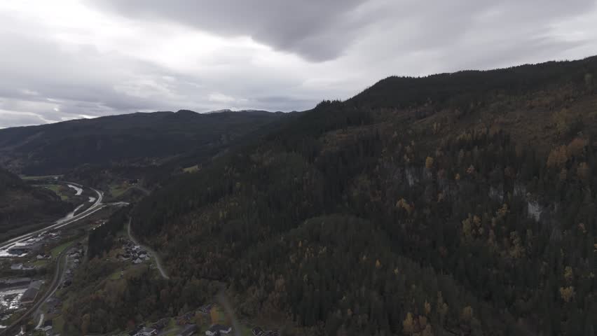 Drone glides along a forested mountain valley, revealing winding roads and a small village below as dark autumn clouds hang low over the hills and river corridor