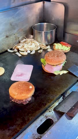 Griddle shot showing multiple burgers, including a ham burger and a beef patty, surrounded by sliced onions and sandwich toppings in a busy kitchen.