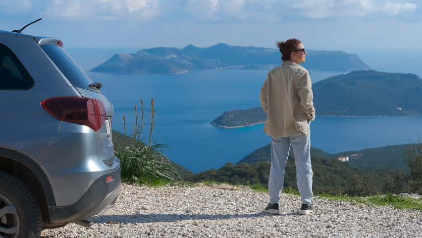 Woman tourist admiring inspiring seascape view from mountain road. Young woman tourist in casual clothing stands next to her car parked on a mountain road, enjoying a breathtaking view of the sea