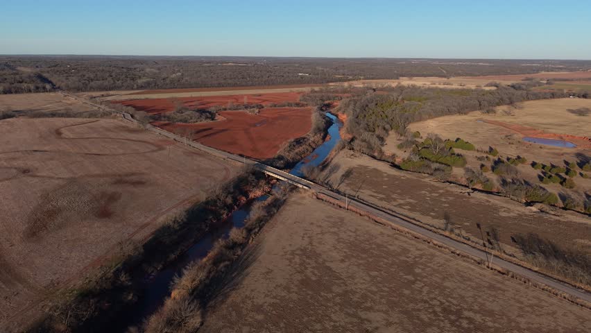 Aerial view of a winding river cutting through red dirt farmland and winter fields in rural Oklahoma under a clear sky.