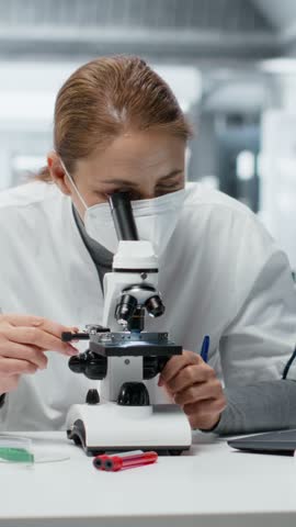 Vertical video Technician wearing surgical mask examining pathology slides under microscope in research center. Researcher using protective medical gear driving medical innovation, doing cellular