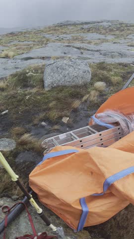 Namsos, Norway – Handheld camera slowly pans across a foggy rocky coastal hilltop as wind-driven rain sweeps over mossy ground, revealing hiking gear beside a small wooden shelter in harsh weather