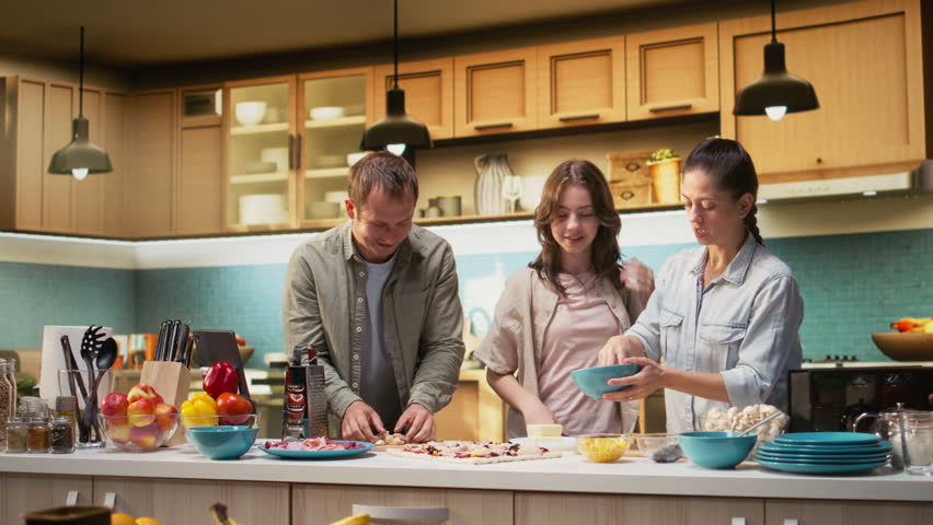 Parents and tween girl laughing while sprinkling mozzarella on pizza in the kitchen, family scene shows bonding time. Culinary creativity and homemade food preparation together. Camera A.