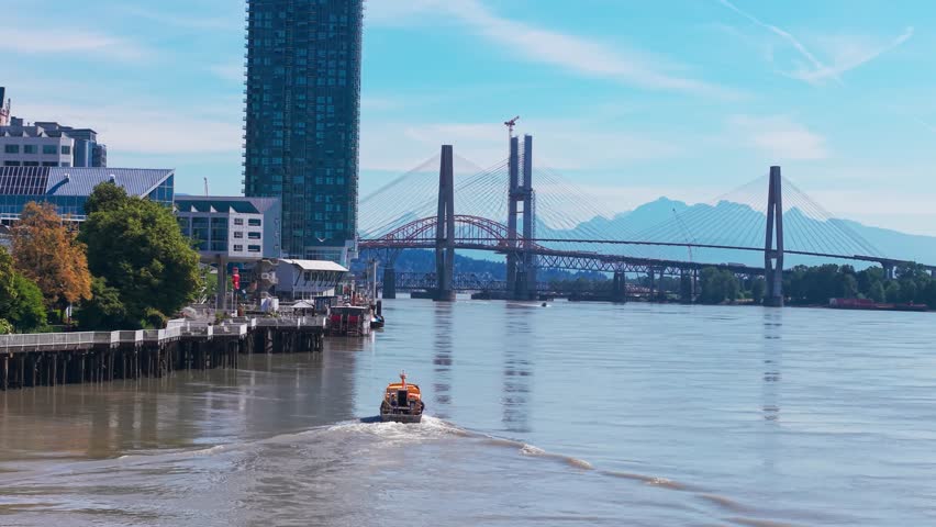 Drone footage of a small boat traveling on the Fraser River toward the Pattullo Bridge in New Westminster, British Columbia, with modern high-rise buildings, waterfront piers, and mountains in the bac